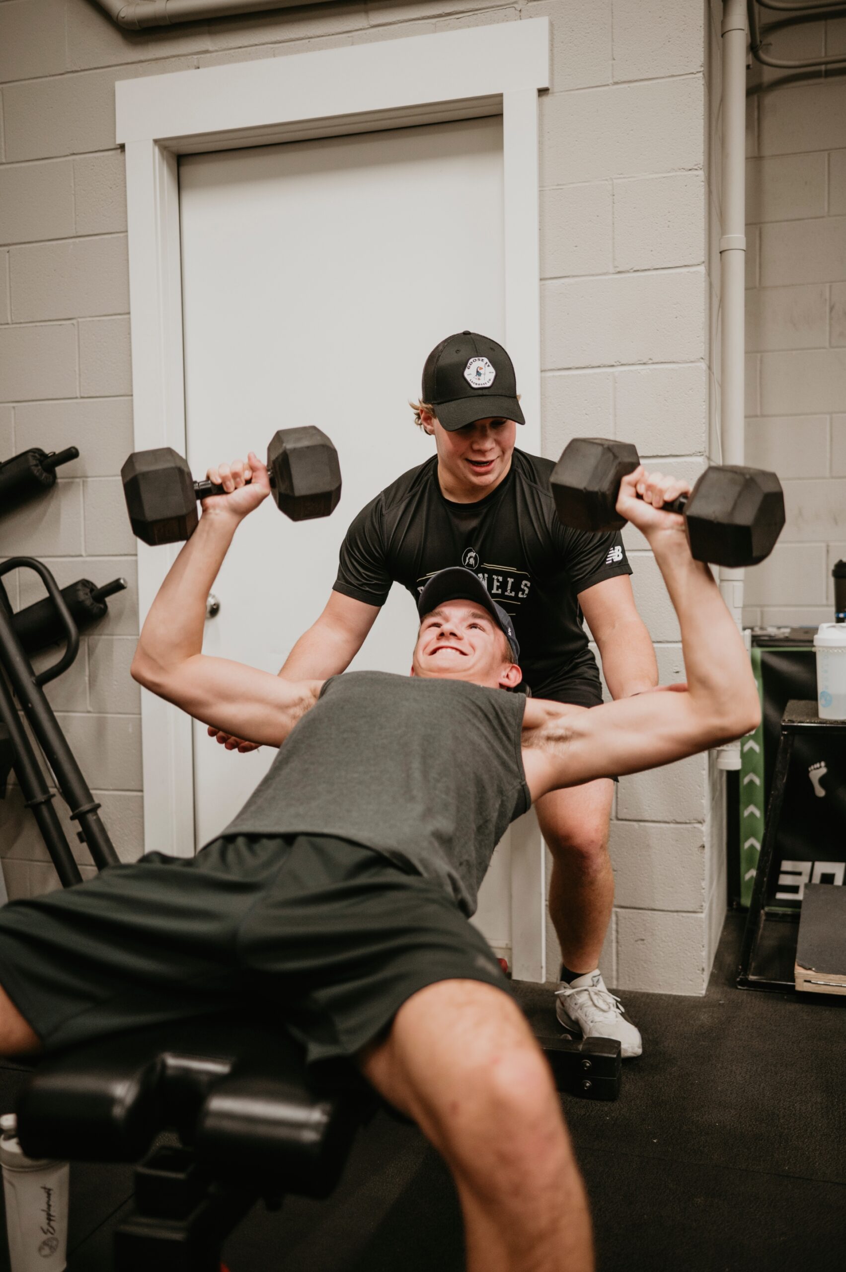 A man performs a dumbbell bench press with a spotter in a gym setting.